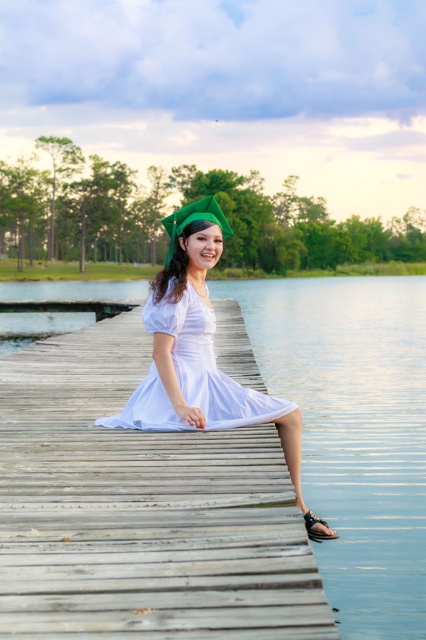 Young woman in white dress and green hat sitting on wooden dock by serene lake at sunset.
