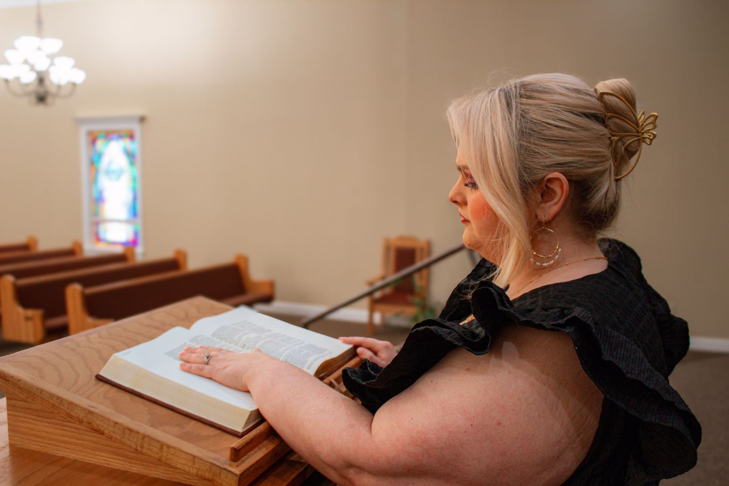 Side view of blonde woman in black dress reading Bible at church pew.