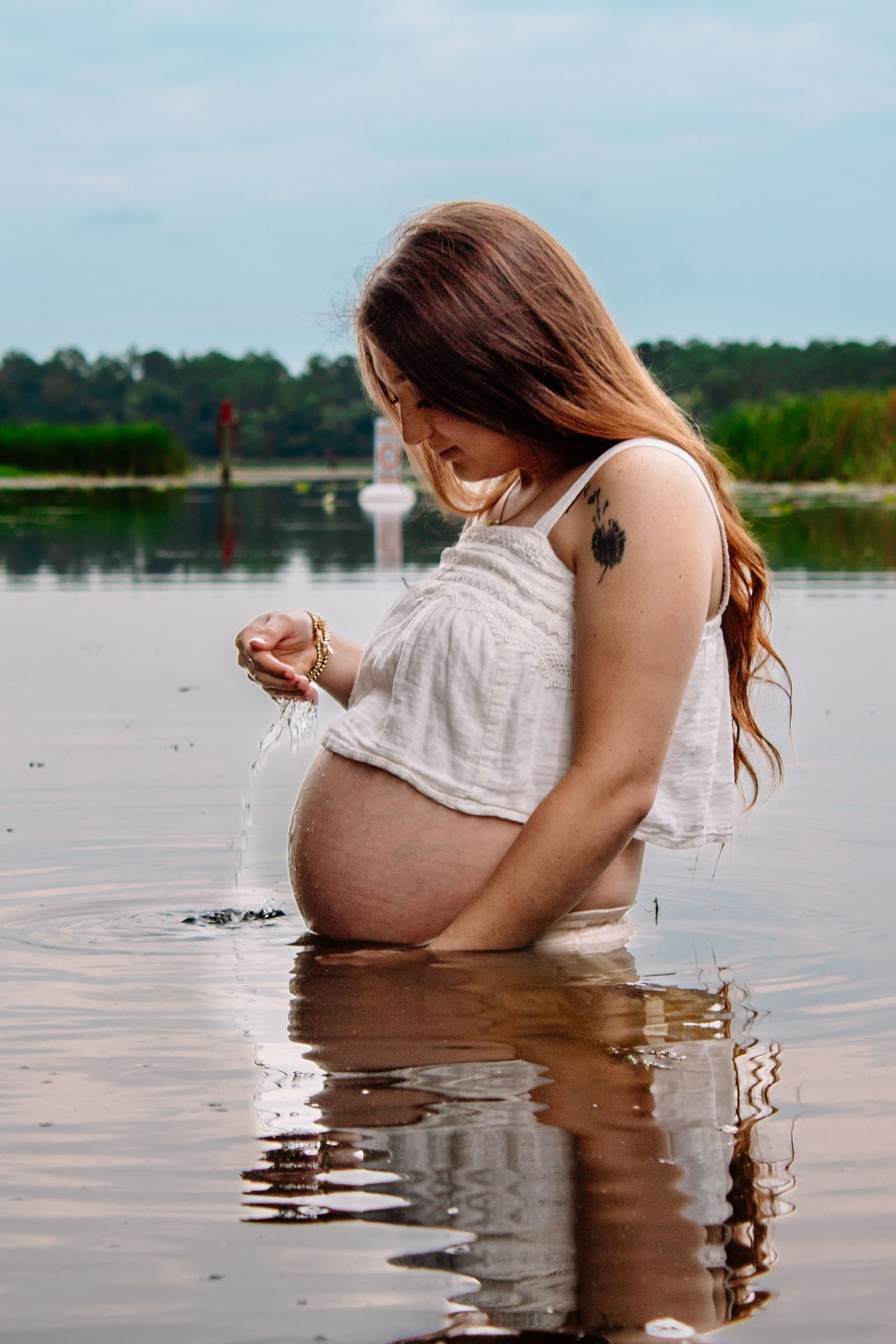 Pregnant woman in white top standing in lake water, letting water drip from her hands over her baby bump.