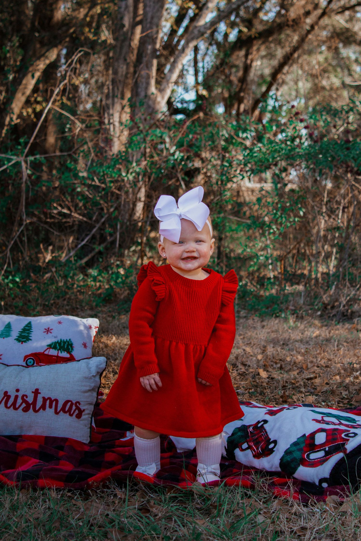 Adorable toddler girl in red dress and white bow standing on Christmas pillows in an outdoor holiday photo.