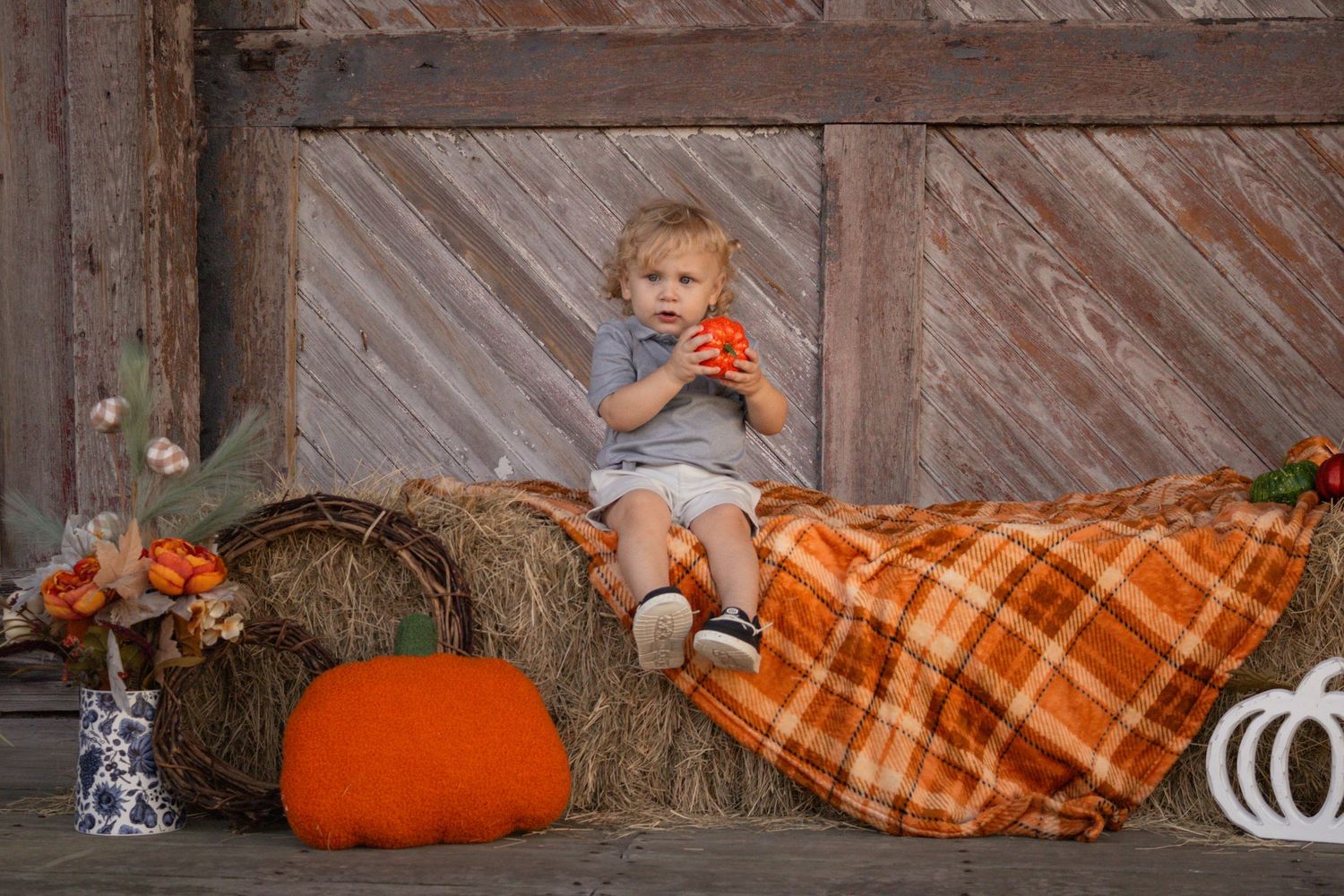 Toddler boy sitting on hay bale with small pumpkin at fall barn photo session.