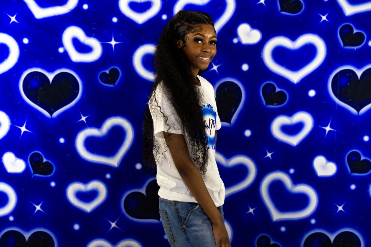 Young Black girl in white tee smiles over shoulder against glowing blue hearts backdrop.