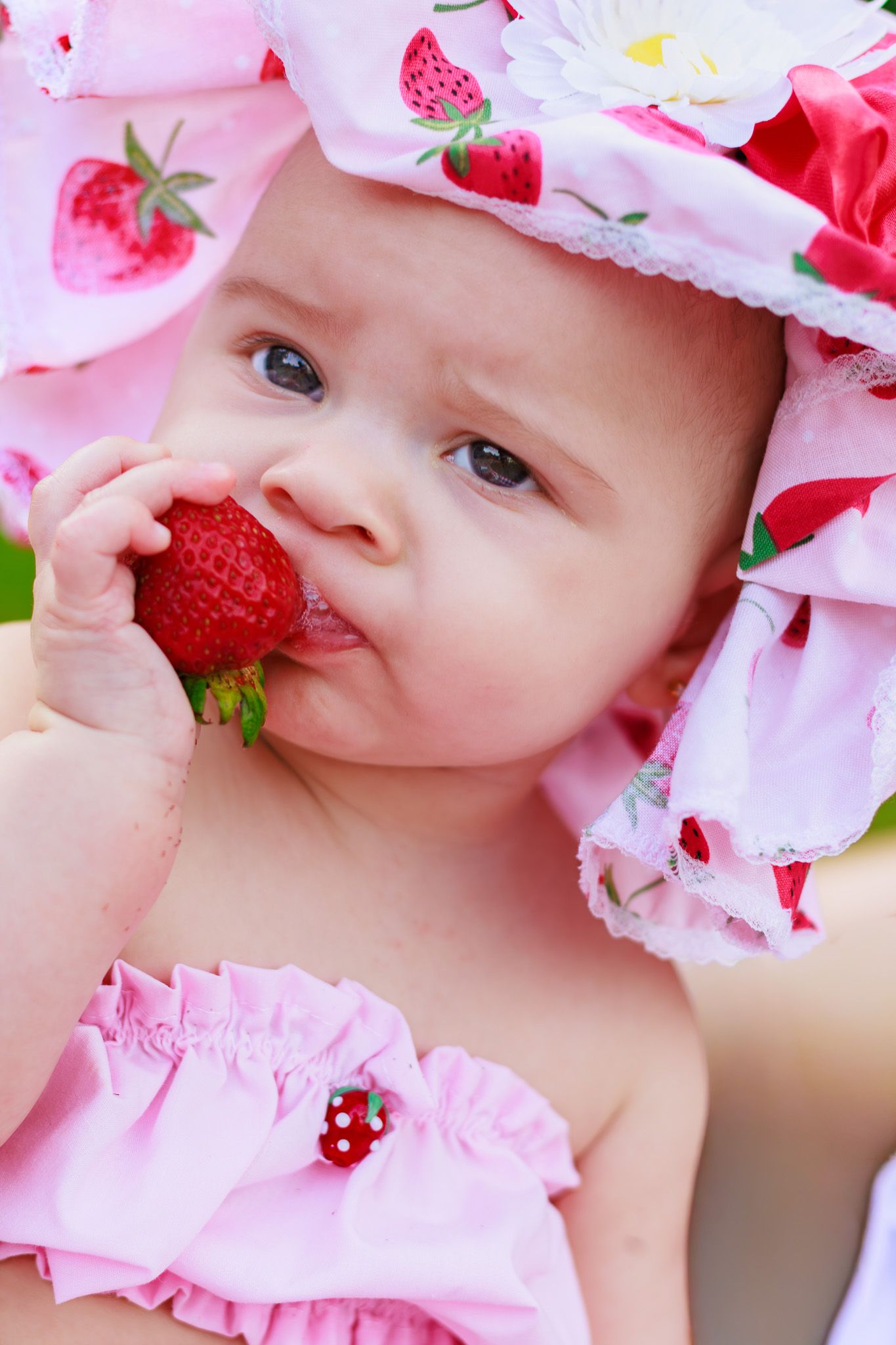 Close-up of sweet baby in pink strawberry sun hat biting into a fresh red strawberry while wearing matching outfit.