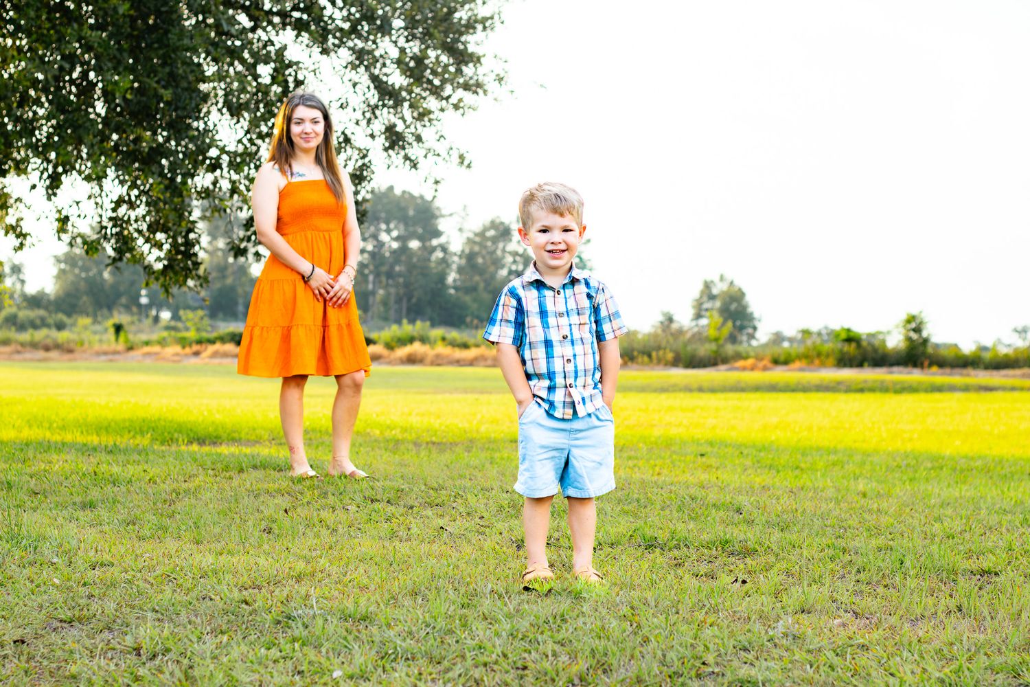 A smiling boy in plaid shirt and shorts stands in green field with a woman in orange dress behind him.