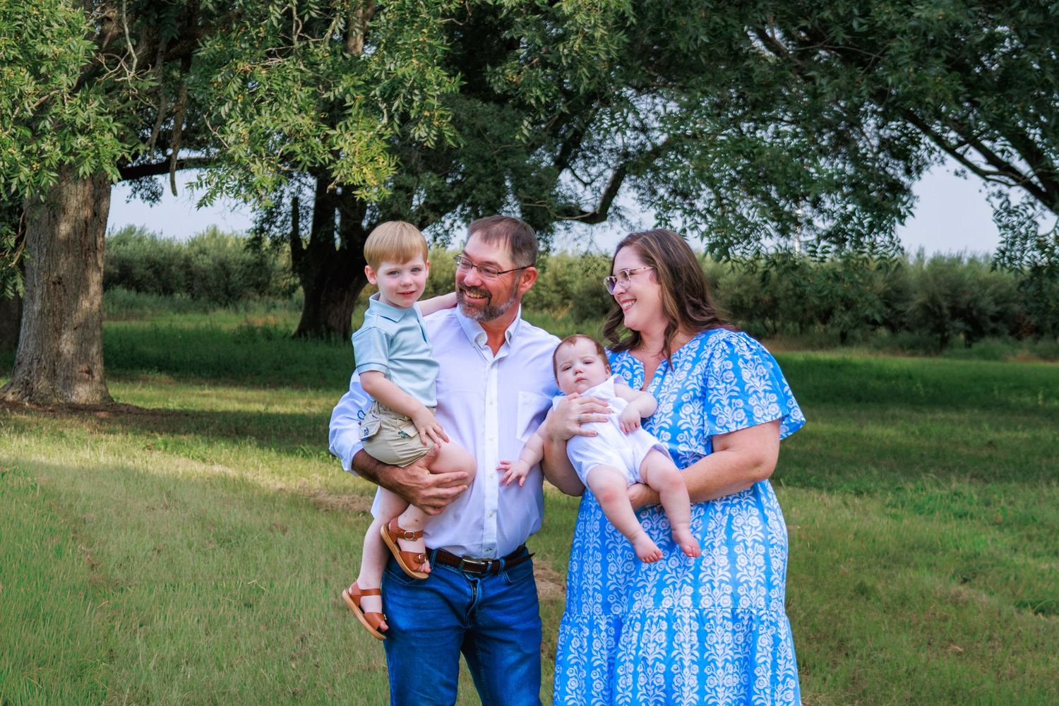 Happy family of four posing outdoors on green grass with large trees, parents holding two young children.
