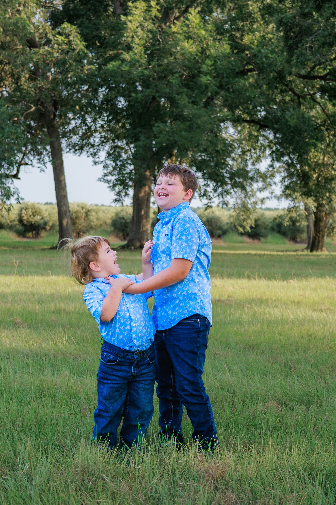 Two boys in matching blue shirts and jeans laugh and play together in a green grassy field with trees.