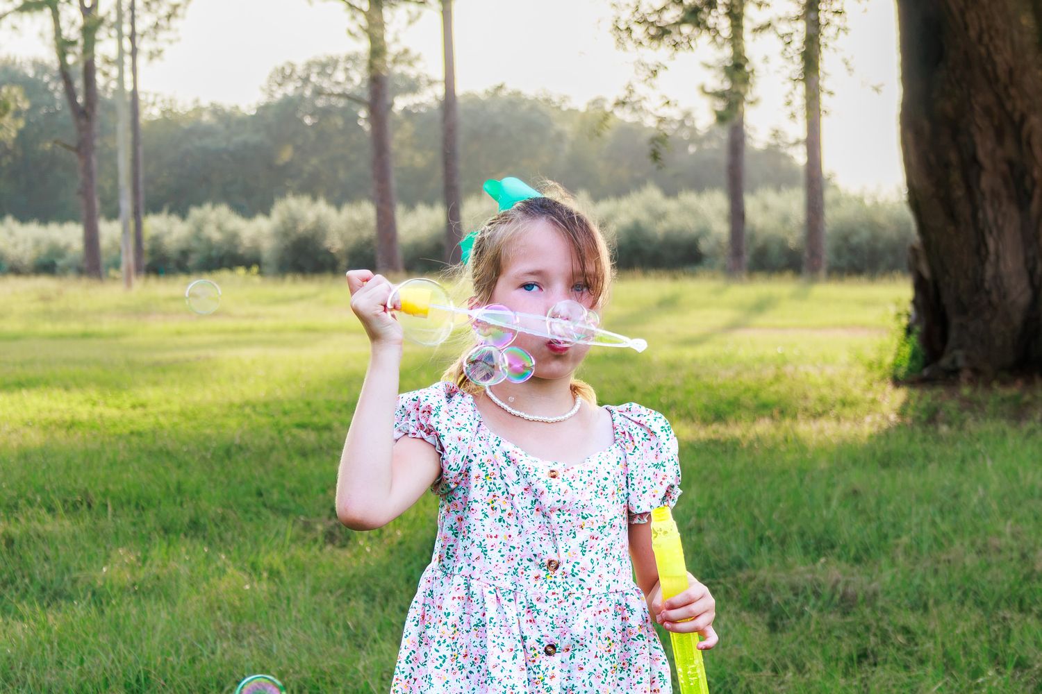 Little girl in floral dress blowing soap bubbles with a wand in a sunny outdoor park setting.