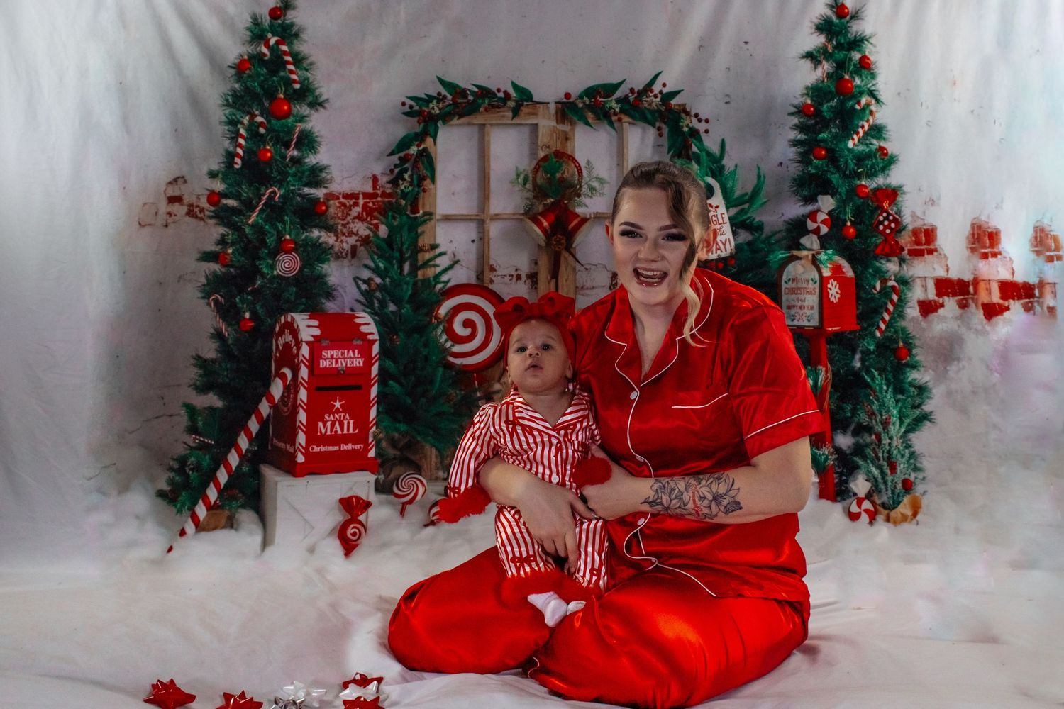 Woman in red pajamas holding newborn in matching outfit in a Christmas-themed candy cane photo studio.