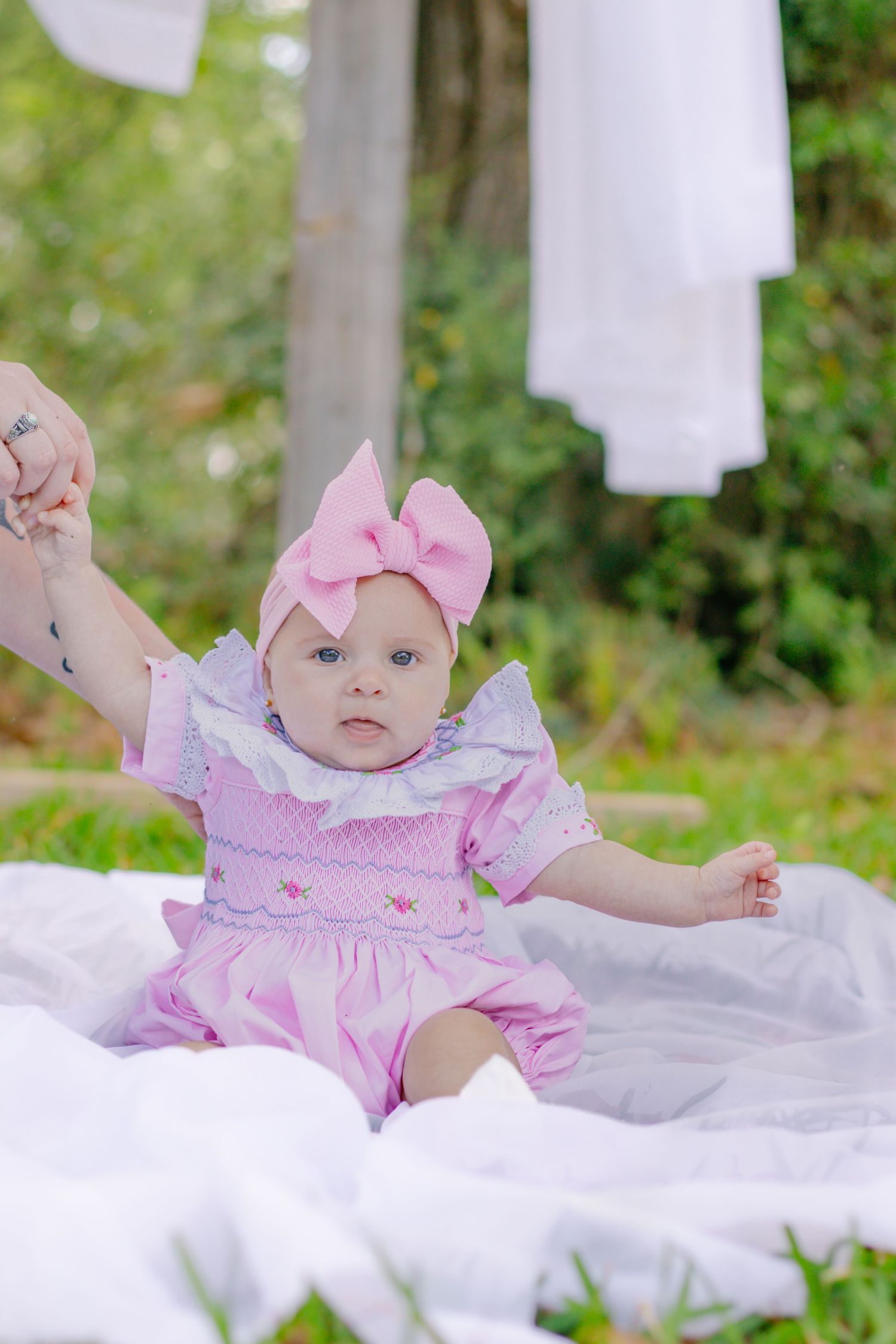 Adorable baby girl in pink smocked romper and pink bow headband sitting on white blanket outdoors.