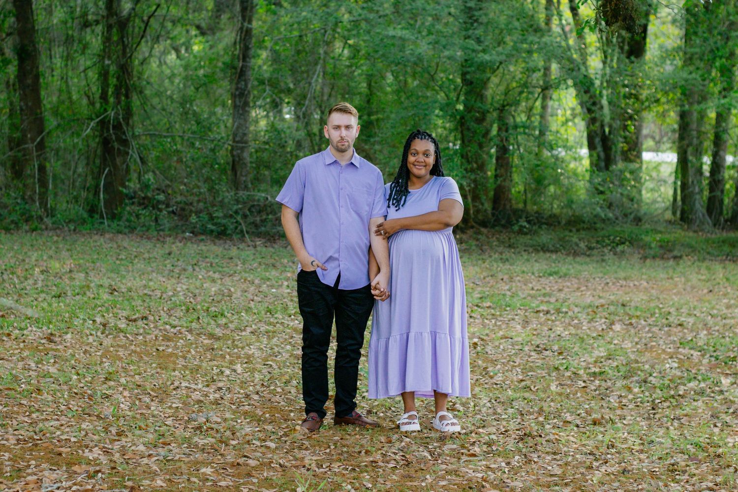 Interracial couple in coordinating lavender outfits stand together in a wooded outdoor setting.