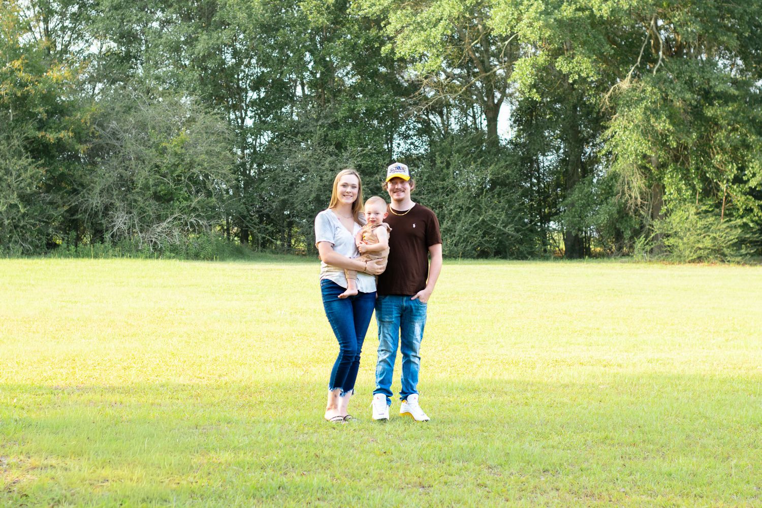 A happy family of three poses outdoors on a green lawn with lush trees in the background on a sunny day.