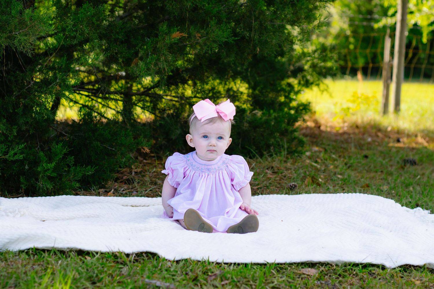 Adorable baby girl wearing pink dress and bow headband sitting on white blanket near green trees.