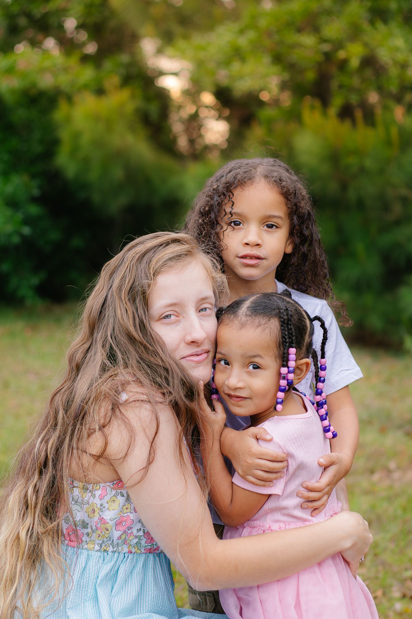 A woman hugging two young girls in a lush green outdoor setting.