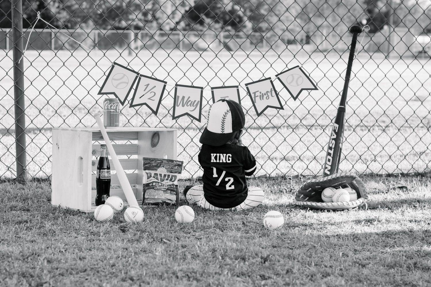 Baby in King jersey sits by baseball field fence with half birthday banner, bats, glove, and balls.