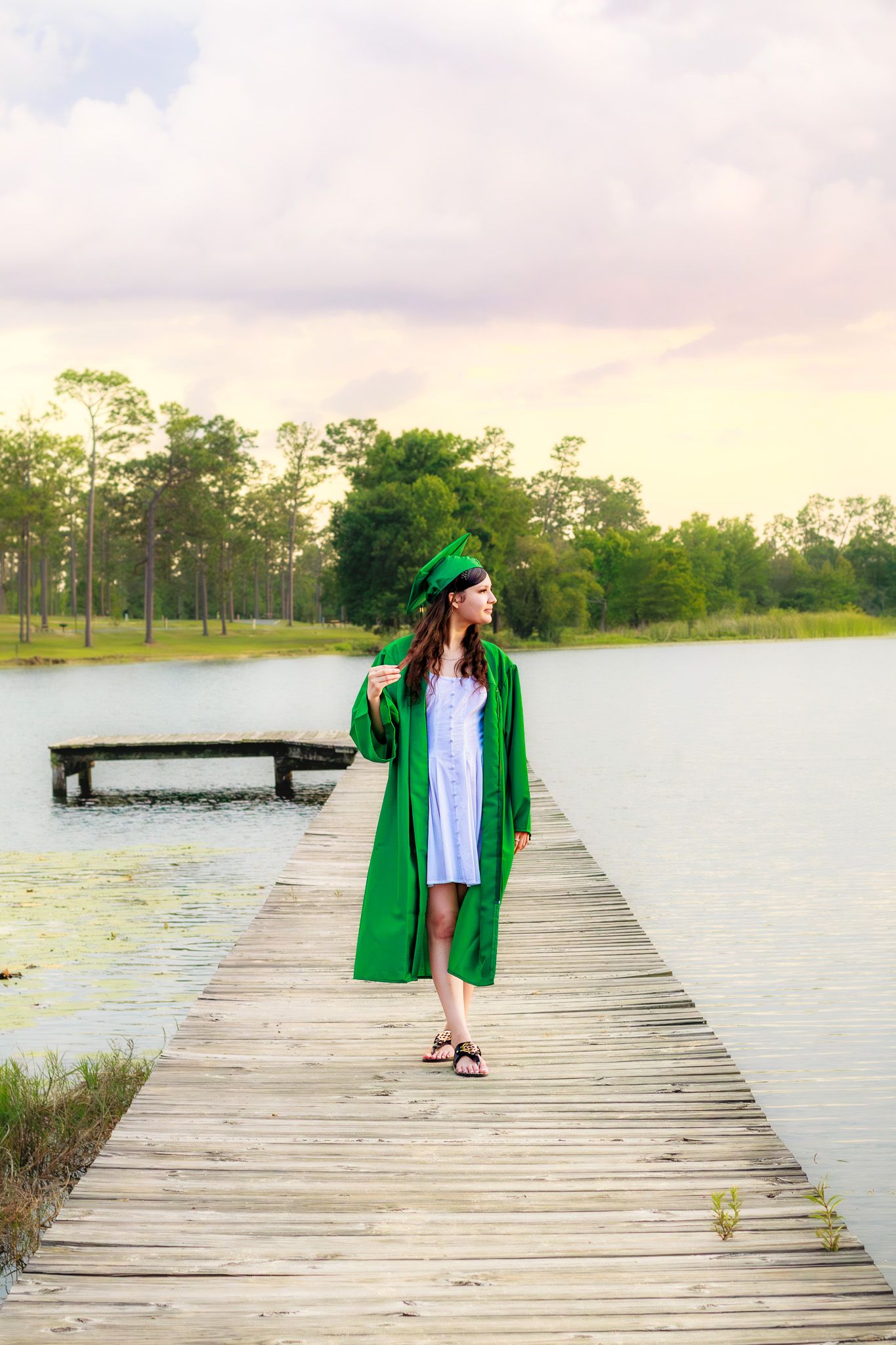 Graduate in green cap and gown standing on wooden dock overlooking calm lake at sunset.