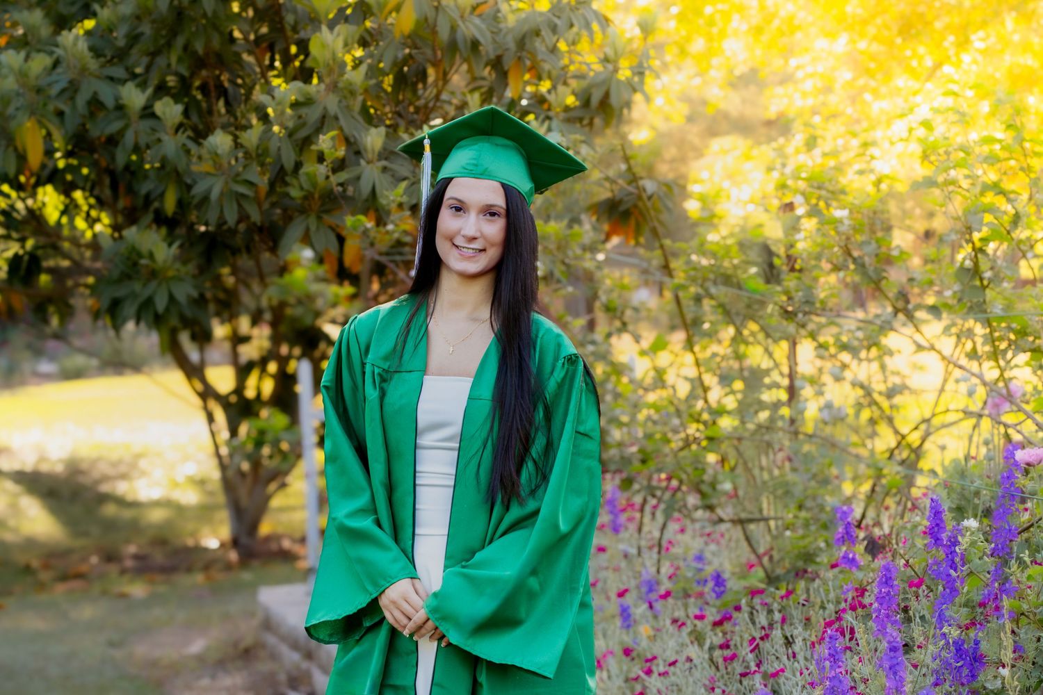 Smiling graduate in green cap and gown posing outdoors among purple flowers and lush green trees.