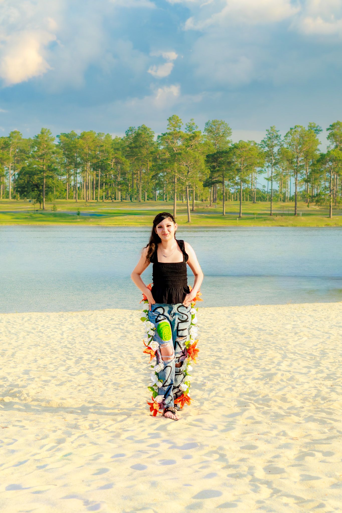 Young woman in colorful printed pants and black top posing on white sand beach with pine trees and lake in background.