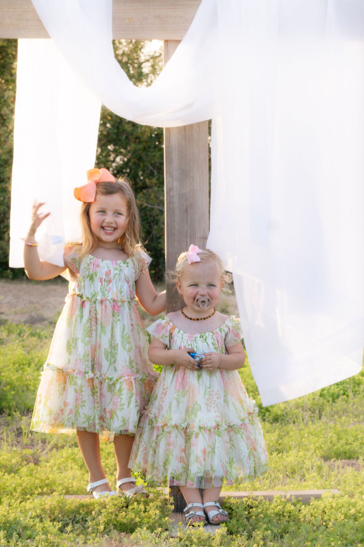Two young sisters in matching floral dresses standing beneath white fabric draped on a wooden cross.