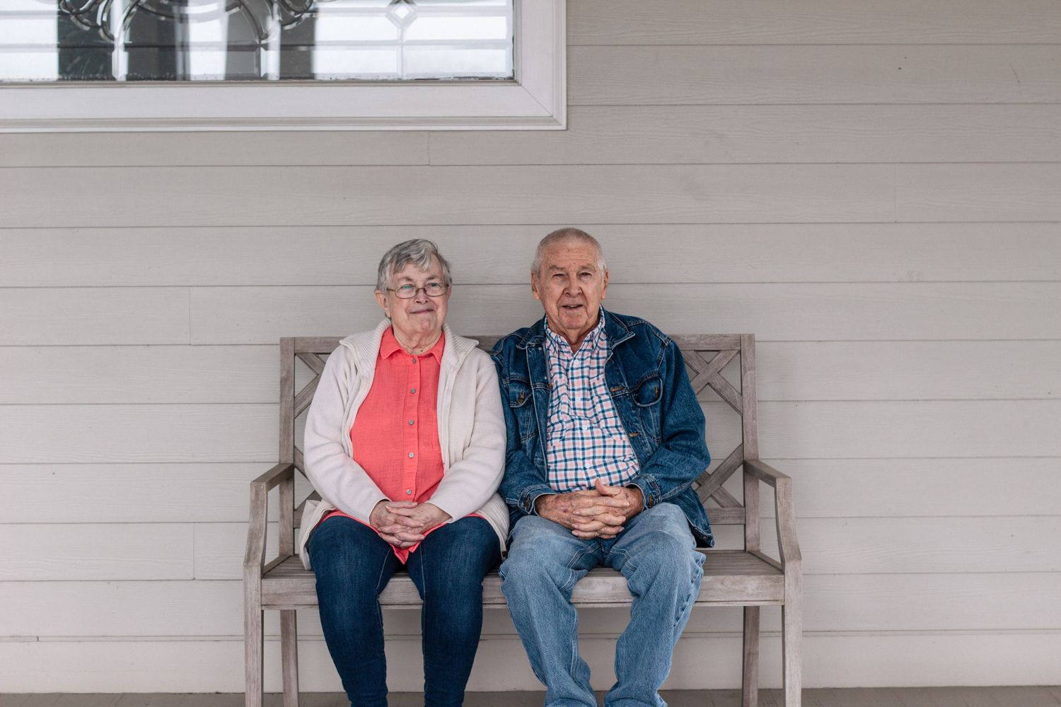 Elderly couple sitting together on a wooden bench on a home porch, smiling warmly at the camera.