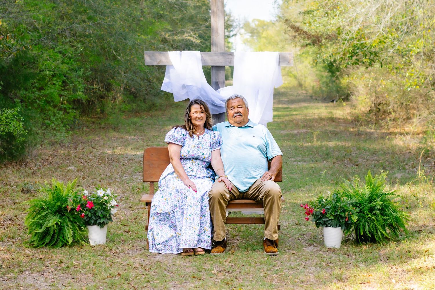 Couple sitting together on a bench in front of a wooden cross with potted ferns and flowers nearby.