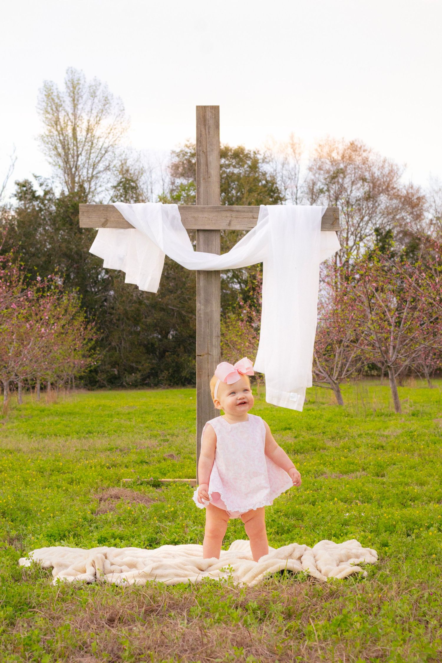Happy baby girl in white dress stands on blanket in front of a wooden cross with white fabric outdoors.