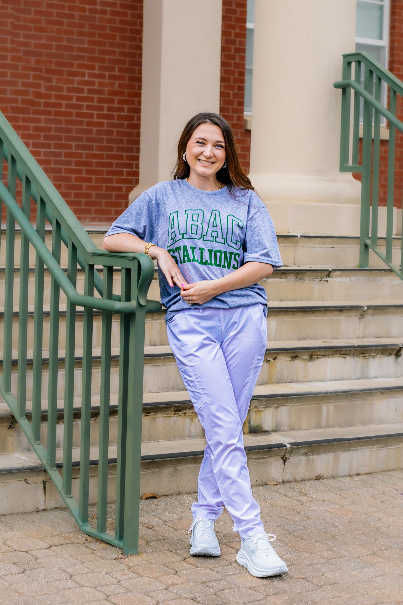 Smiling woman in ABAC Stallions gray tee and lavender joggers leans on green stair railing.