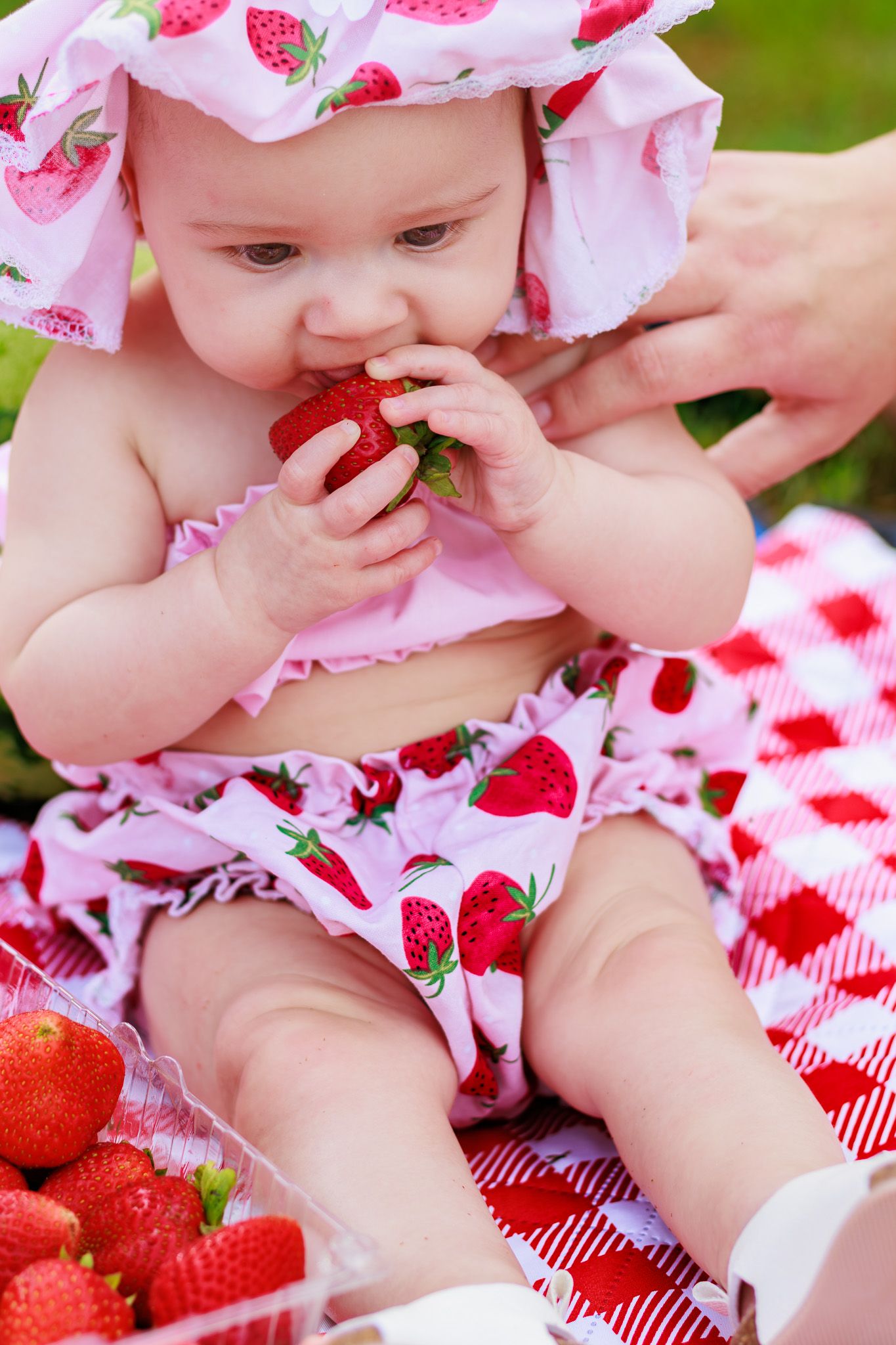 Adorable baby girl in strawberry-print outfit eating fresh strawberry at outdoor picnic on red checkered blanket.