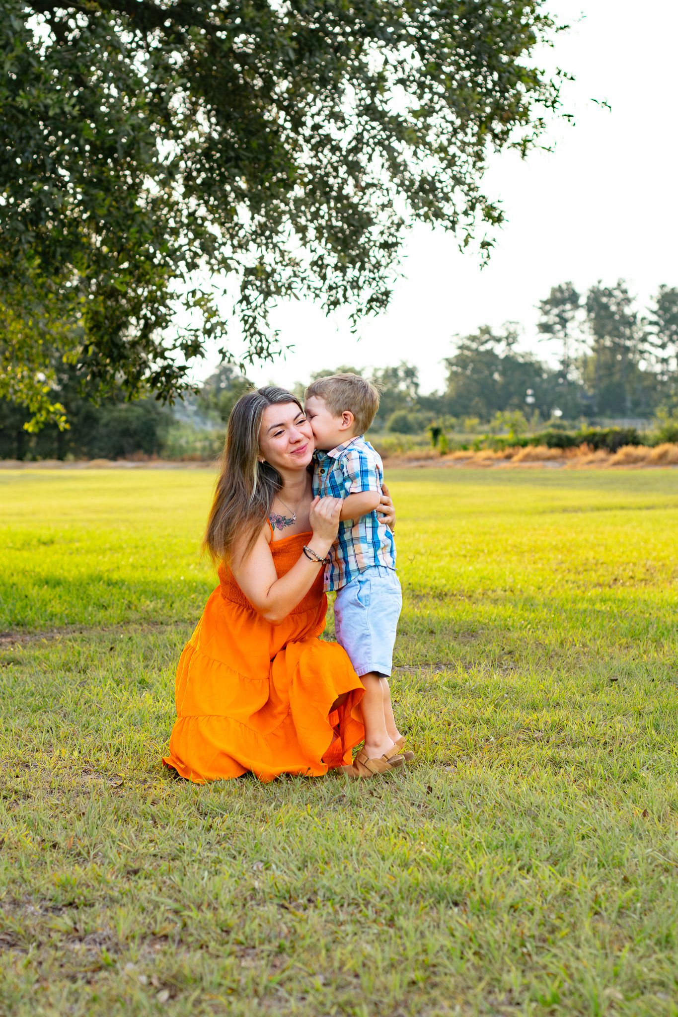 A woman in an orange dress kneels on grass as a young boy in a plaid shirt gives her a kiss outdoors.