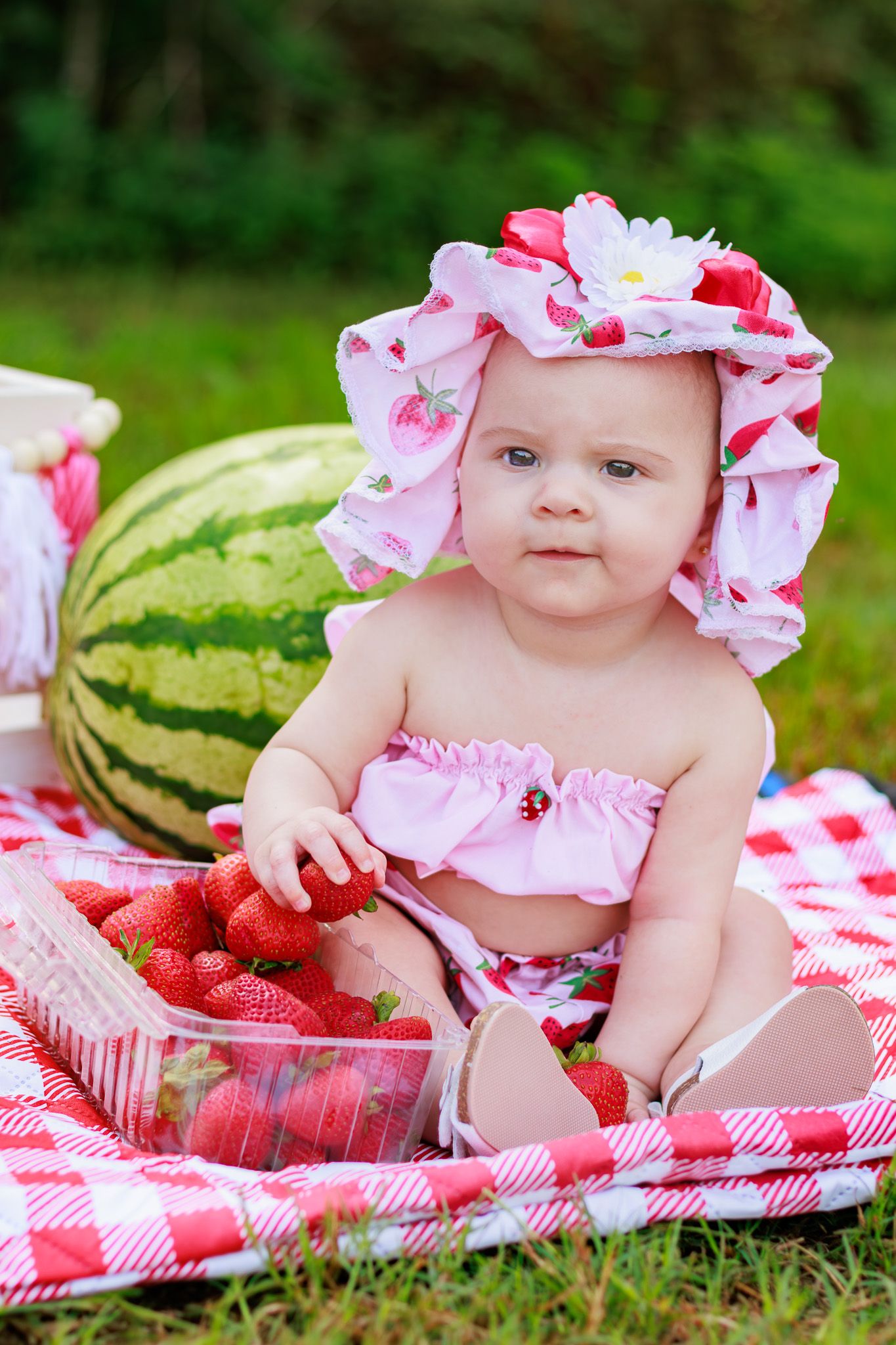 Baby girl in strawberry hat sits on checkered blanket with fresh strawberries and watermelon at summer picnic.