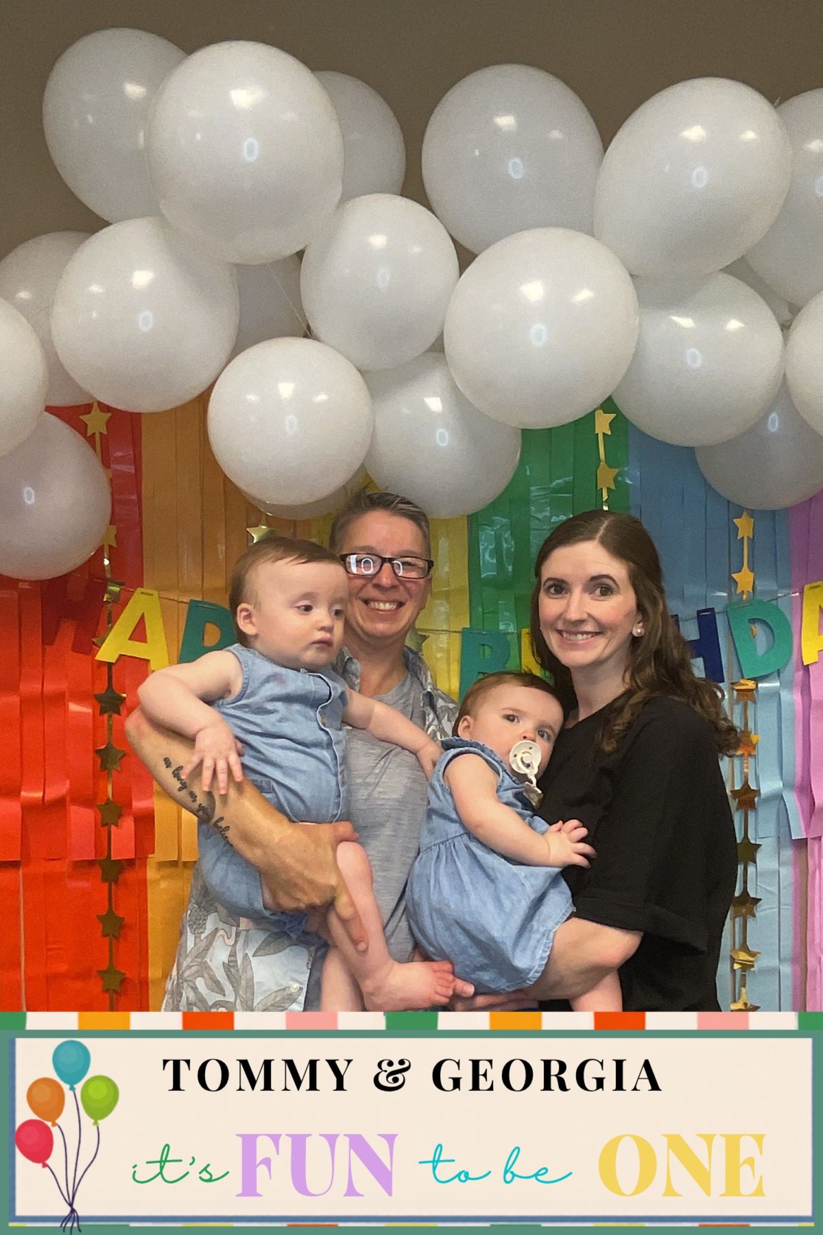 Family photo at Tommy & Georgia's first birthday party with white balloons and rainbow backdrop.