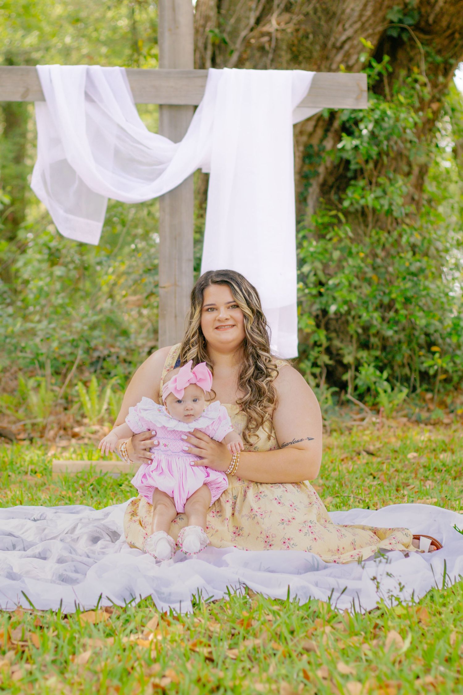 Mother sitting on blanket with baby girl in pink dress before a wooden cross with white fabric.