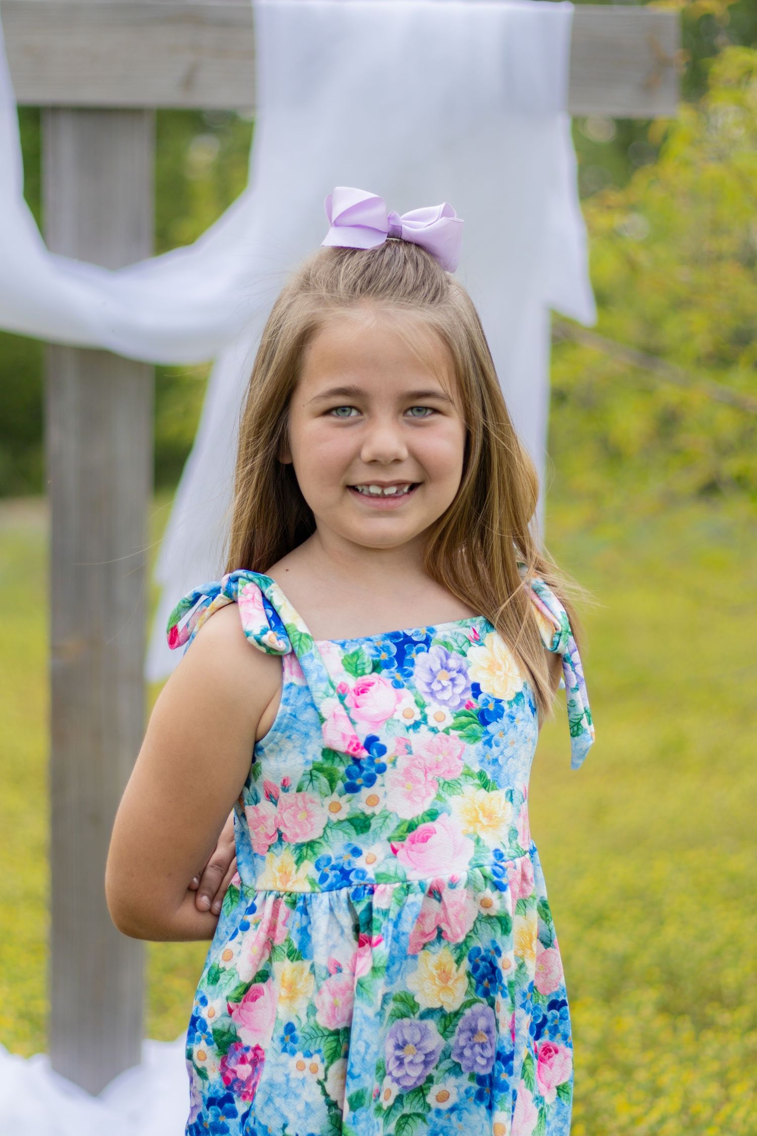 Smiling young girl with purple bow in hair wearing blue floral sundress standing outdoors near a wooden cross.