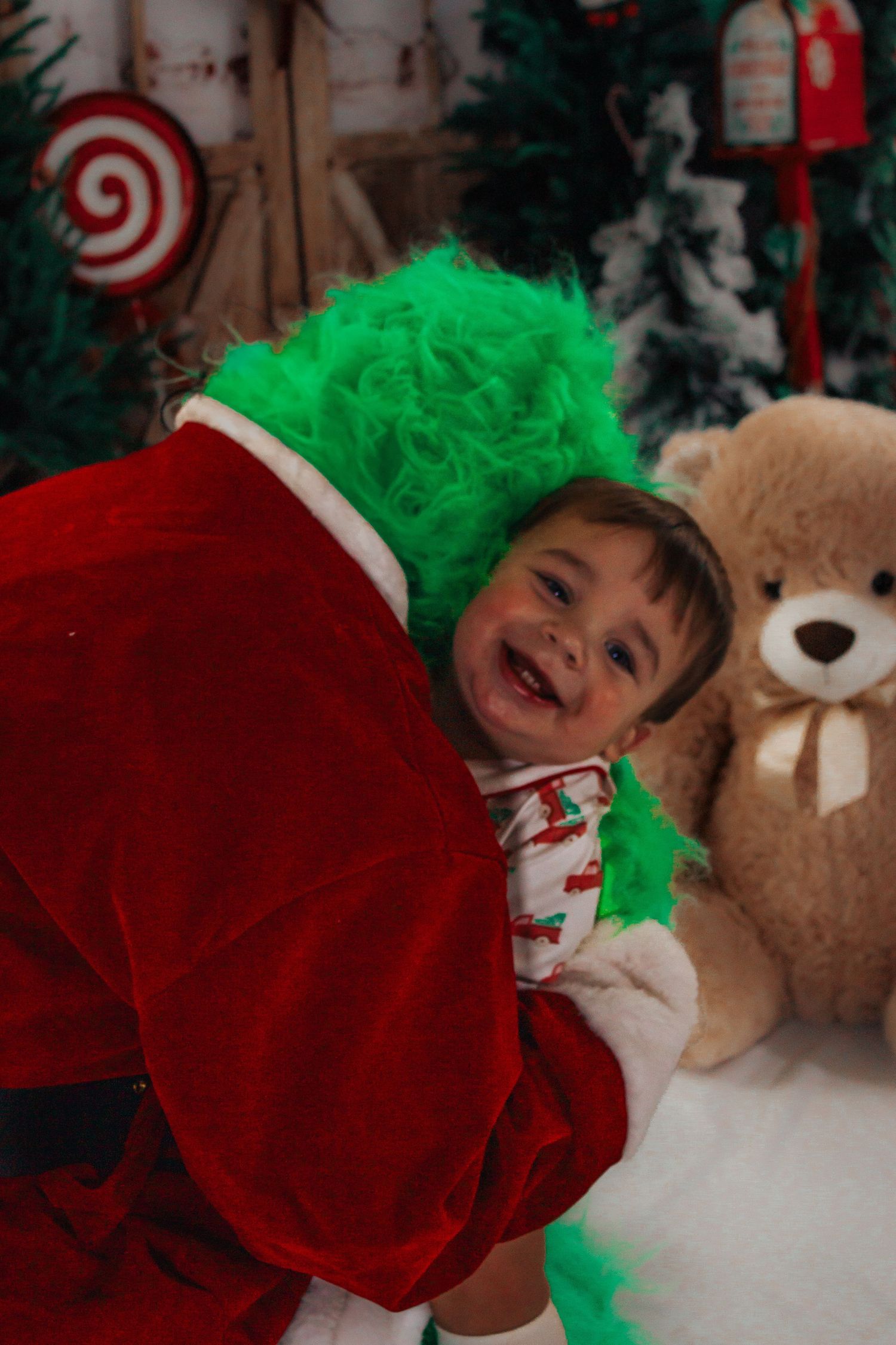 Laughing toddler in Christmas pajamas hugged by someone dressed as the Grinch in a festive holiday photo studio.
