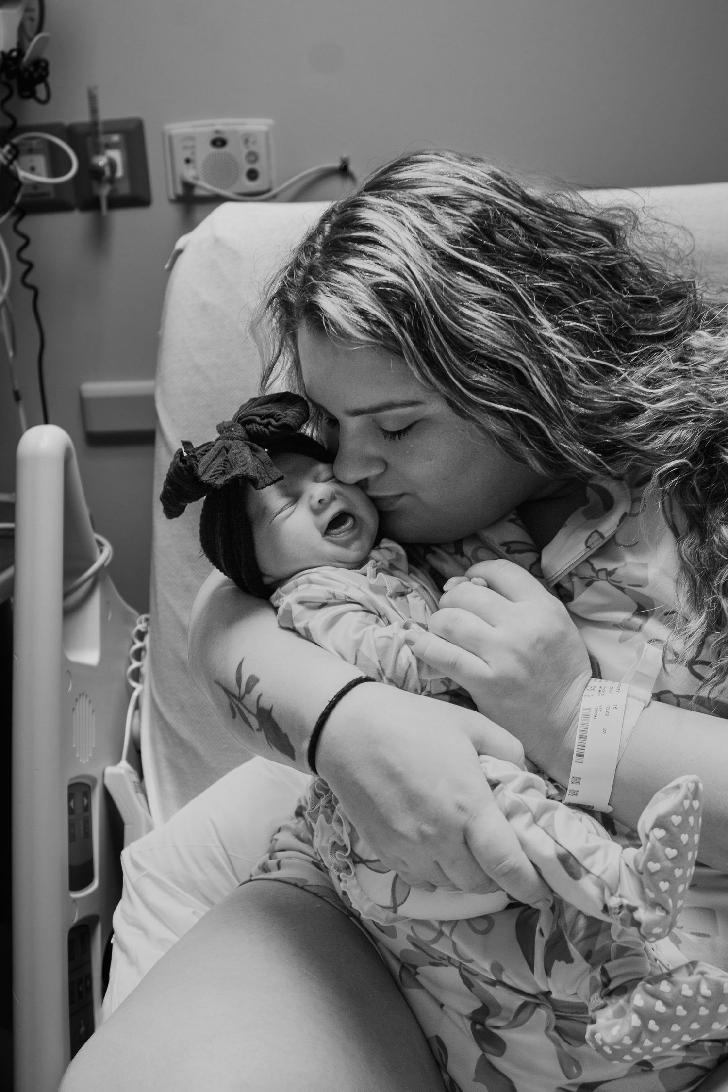 Black and white photo of mother kissing her smiling newborn baby in hospital bed.
