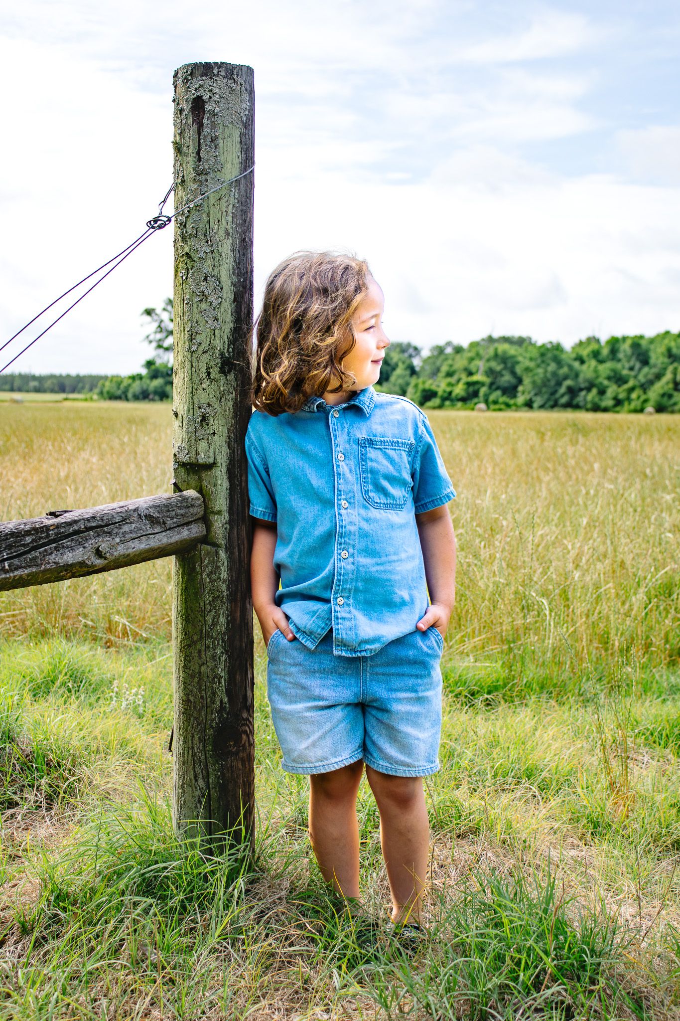 Curly-haired boy in blue denim outfit leaning against wooden fence post in a rural grassy field.