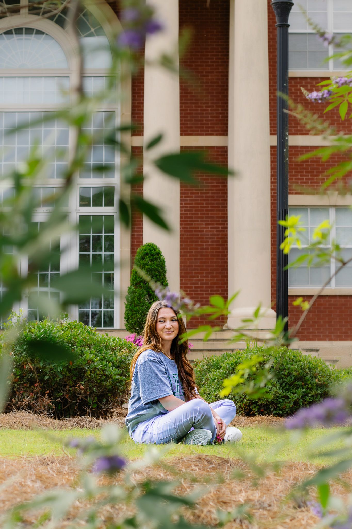 Young woman sitting cross-legged on grass in front of a brick building, smiling through green foliage.