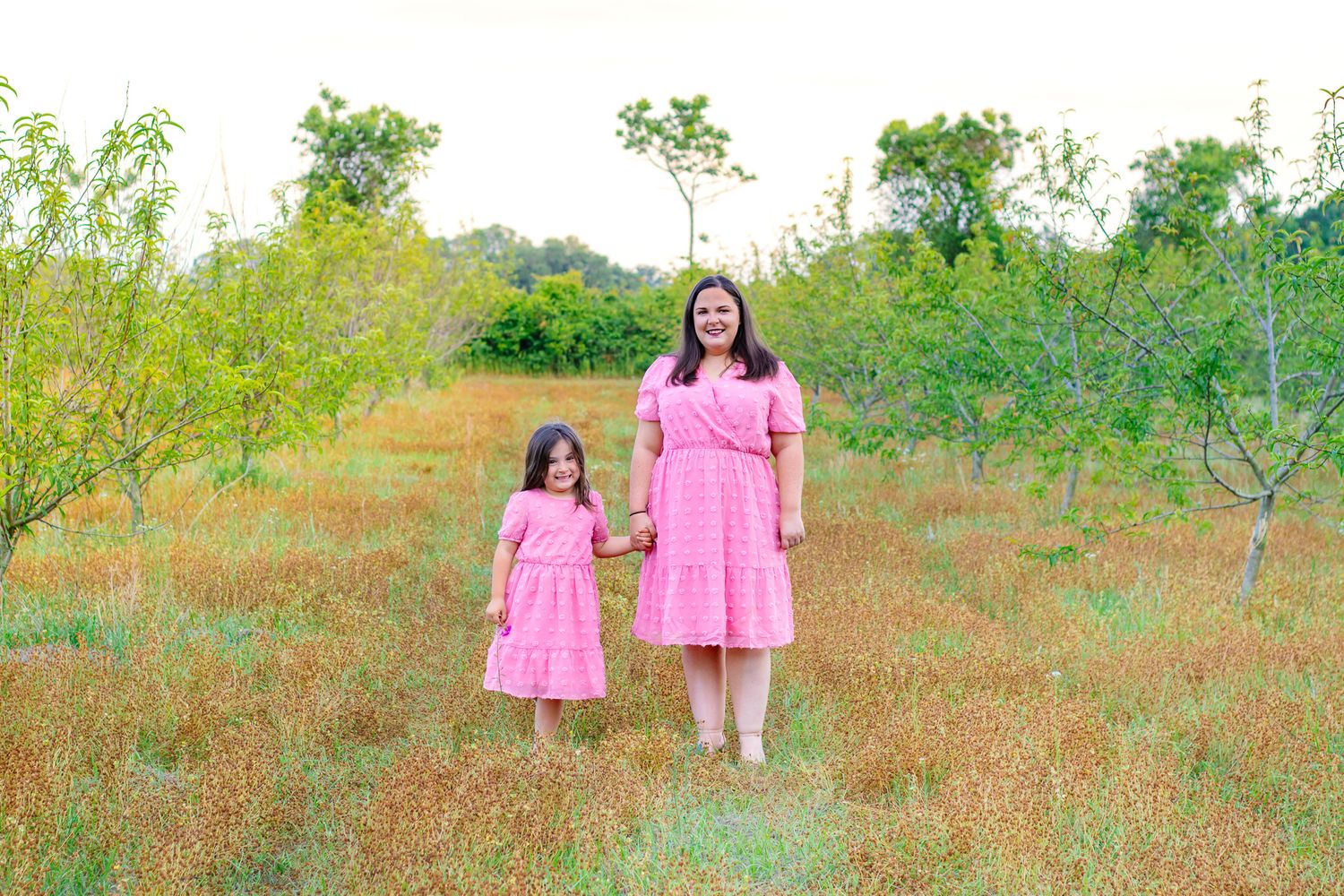 Mother and daughter in matching pink dresses holding hands in a sunny meadow.