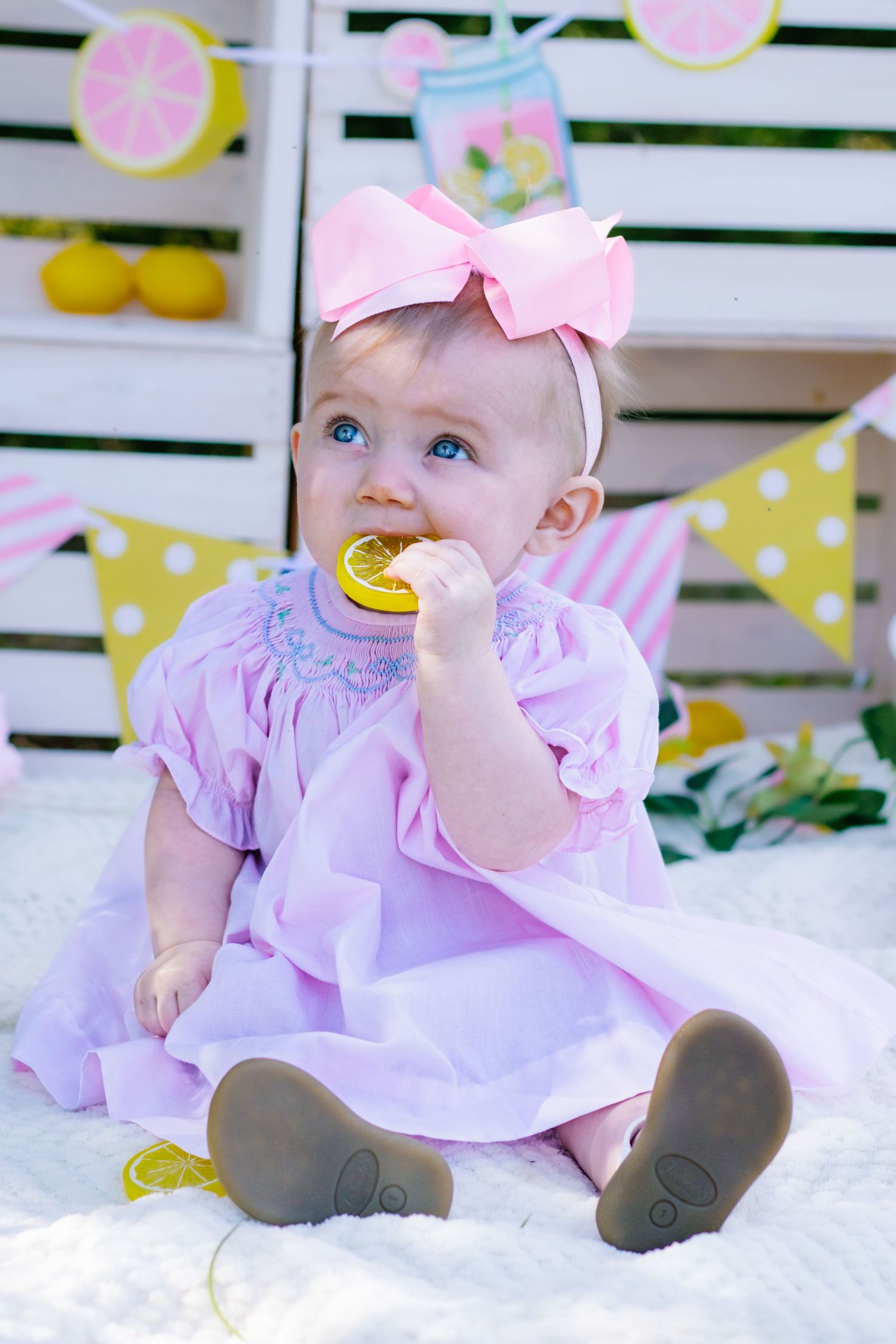 Baby girl in pink smocked dress chewing lemon toy at colorful lemon-themed birthday backdrop.