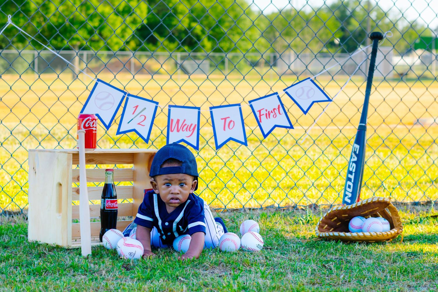 Baby boy in baseball uniform poses on grass with bat, glove, balls, and a Way To Go banner.