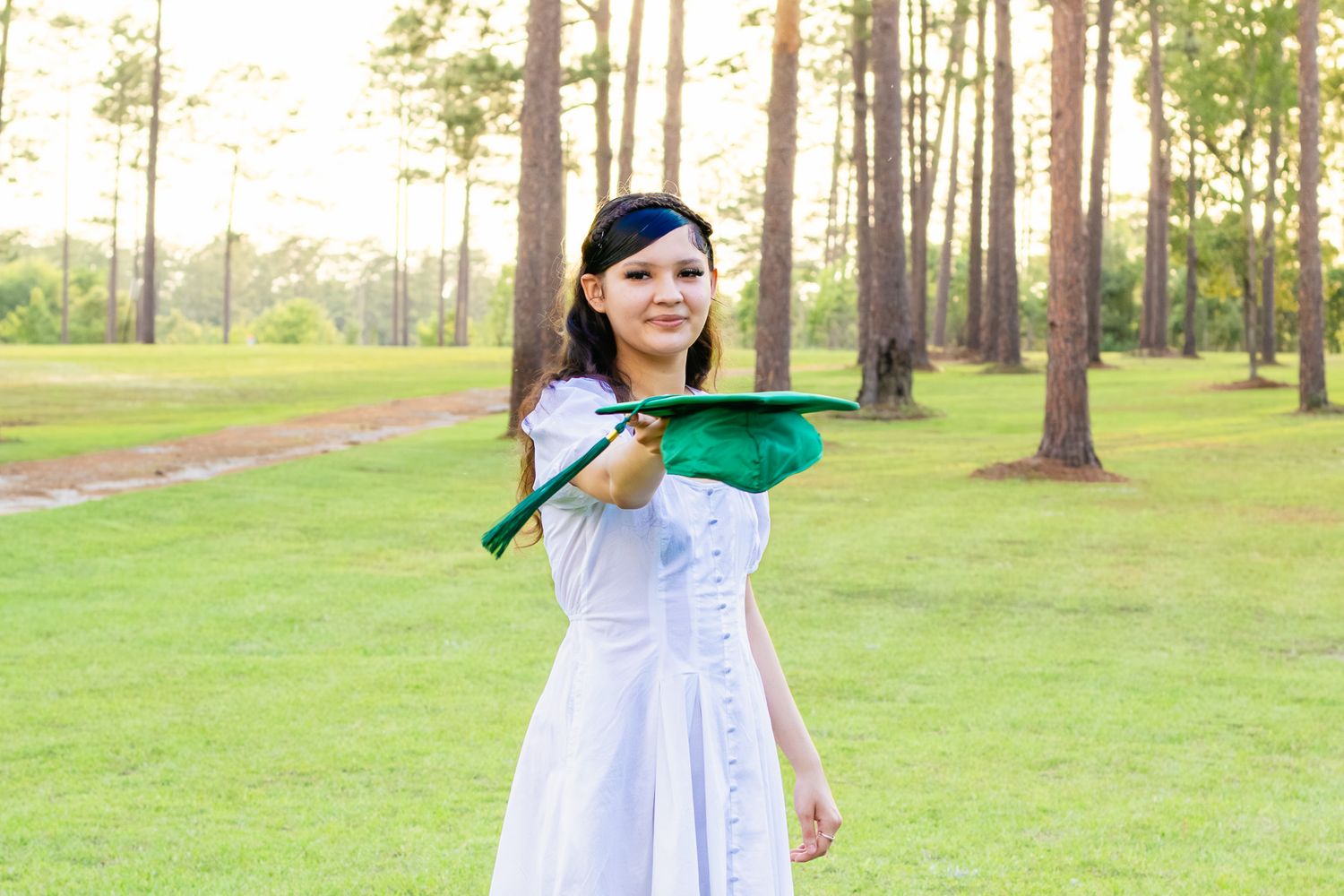 Young woman in white dress holding green graduation cap outdoors among tall pine trees on a sunny day.
