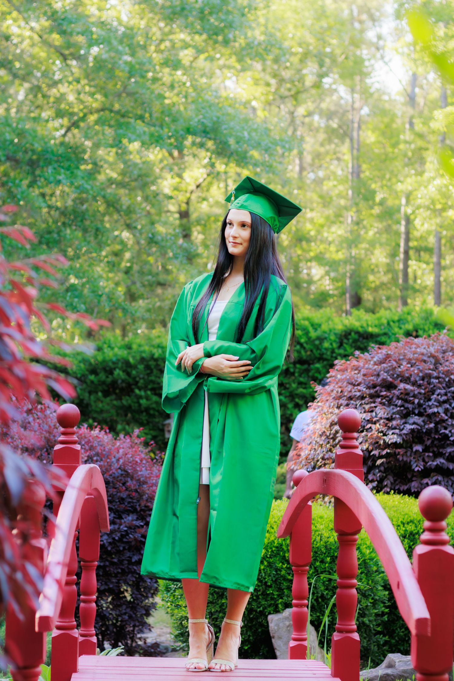 Young woman in green graduation cap and gown standing on a red bridge surrounded by lush greenery.