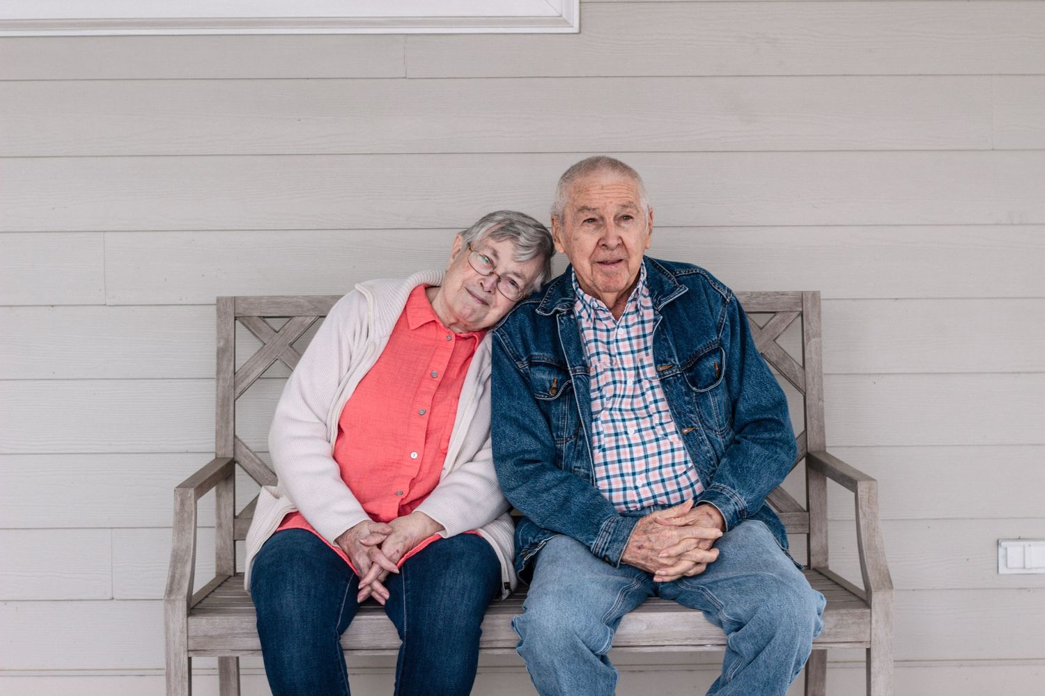 Senior couple sitting close together on a bench, posing for a portrait on a porch.