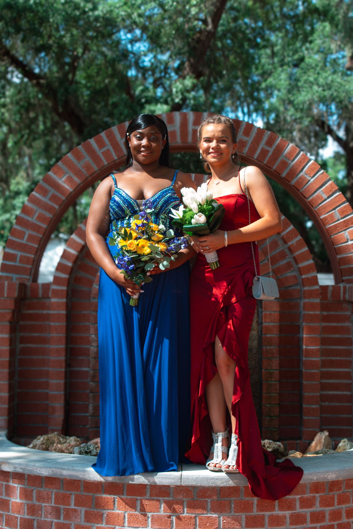 Two smiling young women in blue and red formal gowns holding bouquets under a brick arch outdoors.