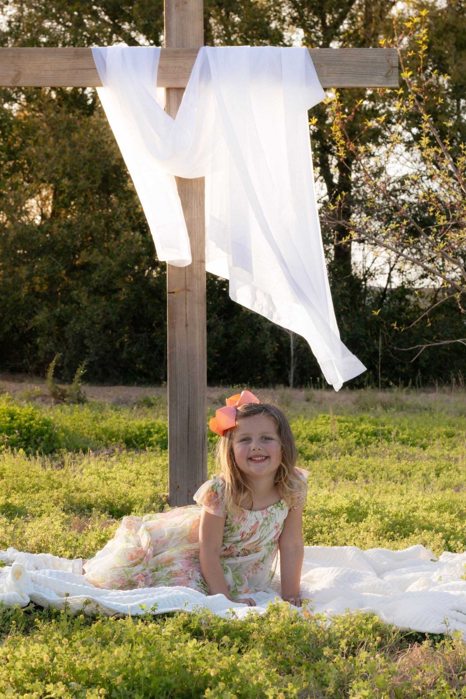 Young girl in floral dress smiling while sitting on white blanket beneath a wooden cross outdoors.