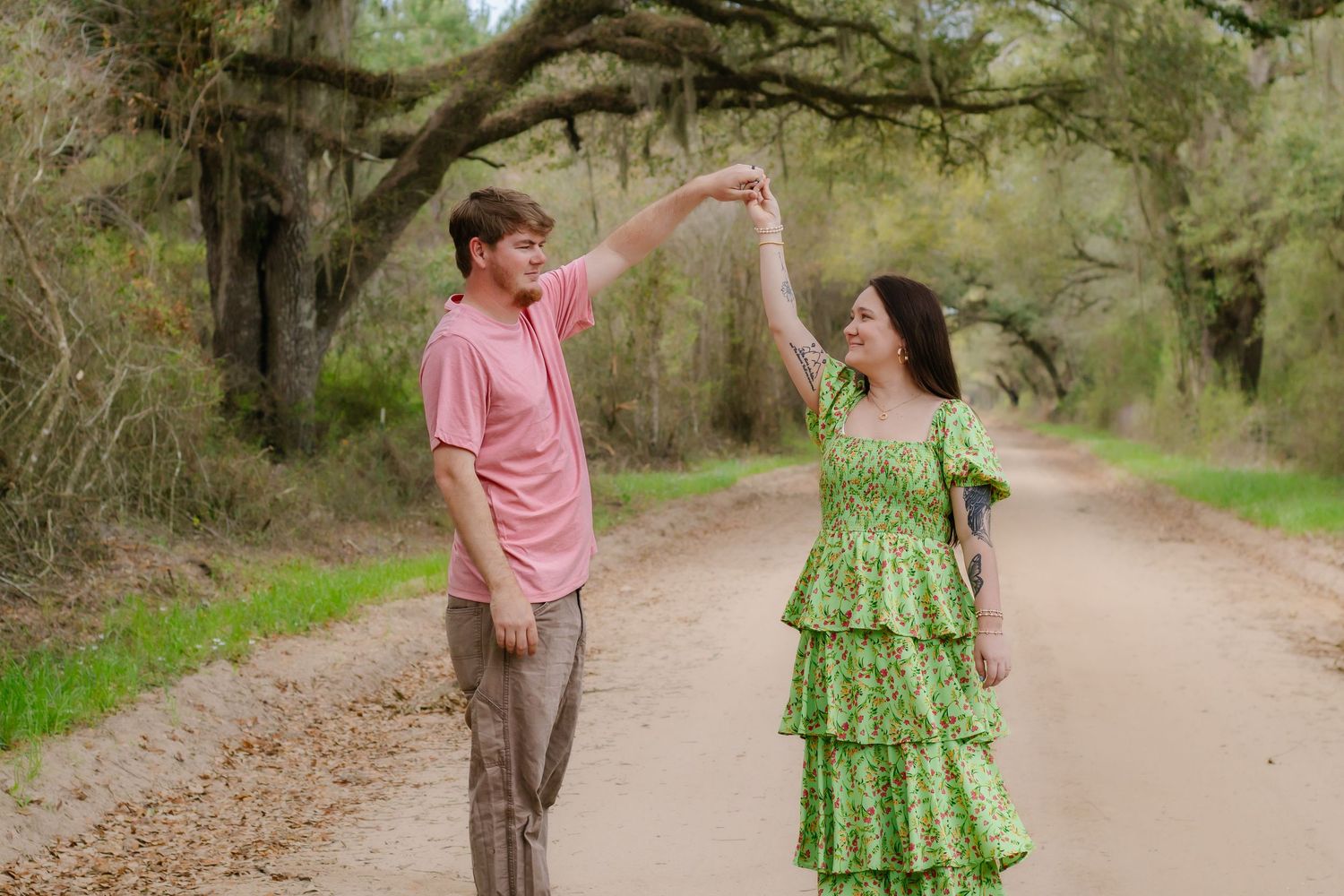 Couple dancing on a scenic dirt road surrounded by lush green trees, man in pink shirt twirling woman in green floral dress.