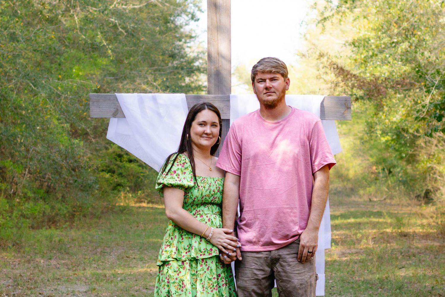 Young couple standing together before a wooden cross outdoors, woman in green dress, man in pink shirt.