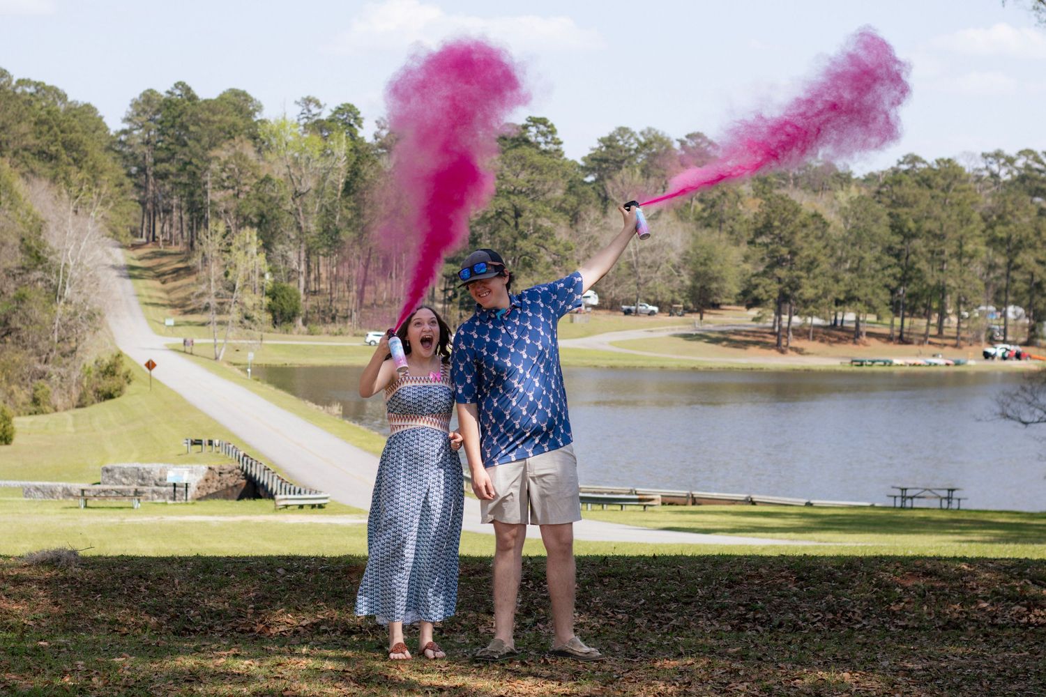 Couple celebrates gender reveal with pink smoke bombs in a scenic park by a lake.