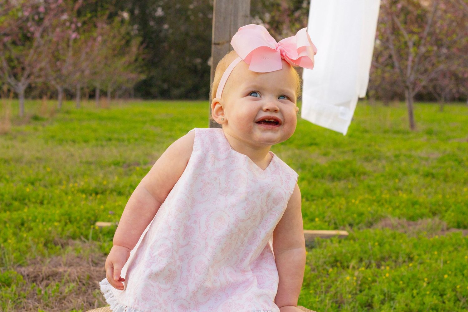 Cheerful baby girl wearing pink bow headband and white dress sits outdoors near a wooden cross in spring.