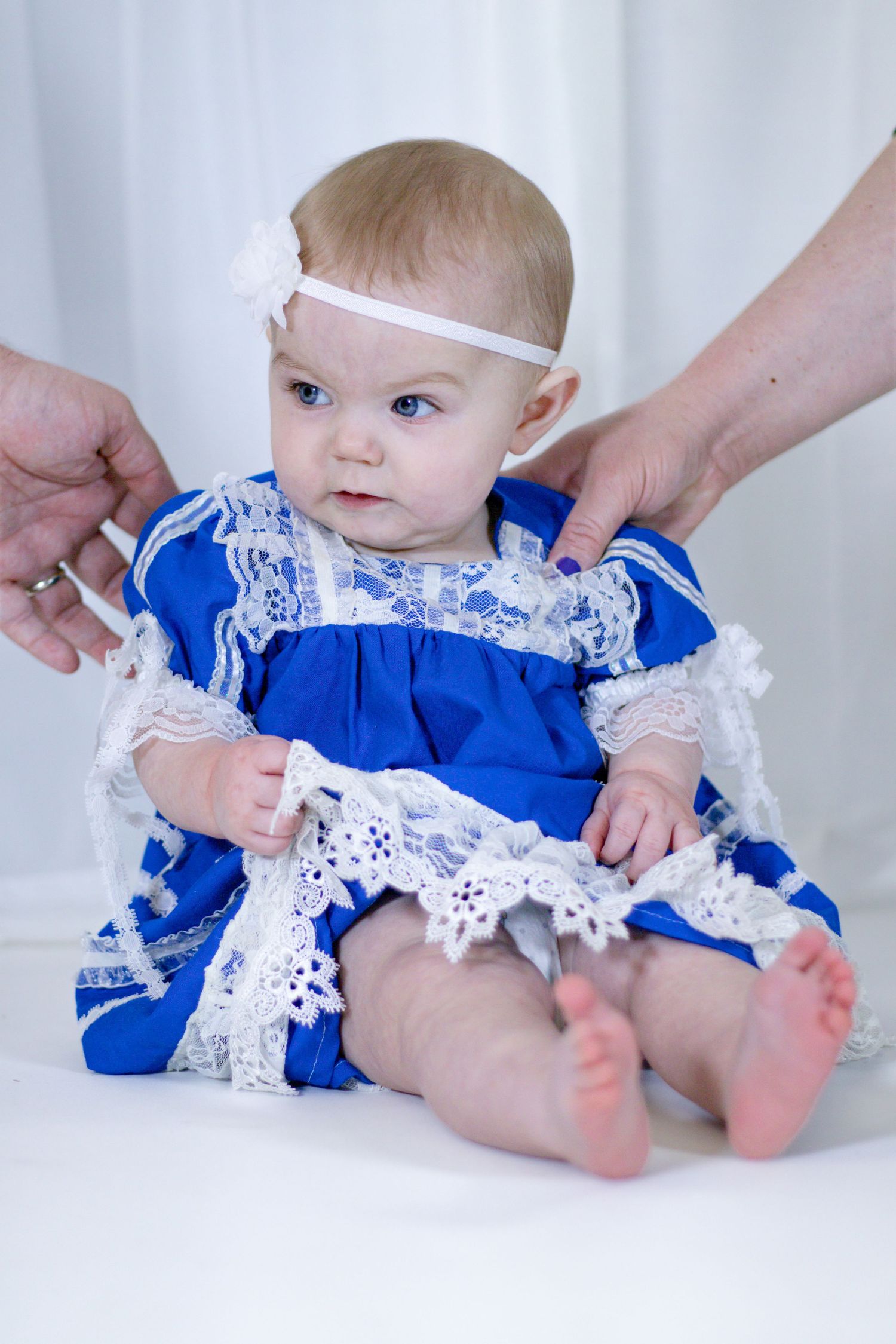 Adorable baby girl in blue lace dress sitting upright with adult hands adjusting her white headband.