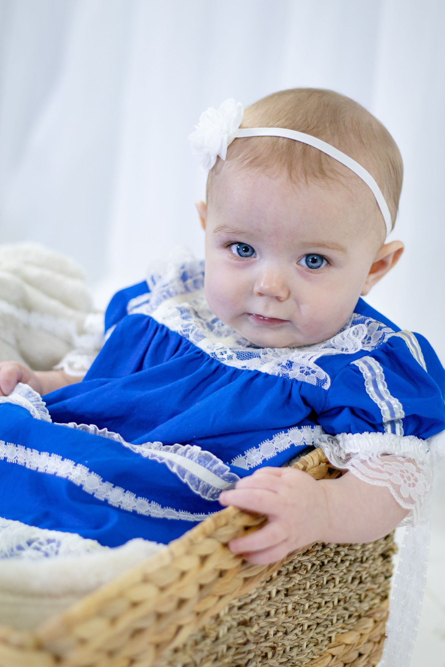 A cute baby girl in a bright blue lace dress with white headband sits in a woven wicker basket.